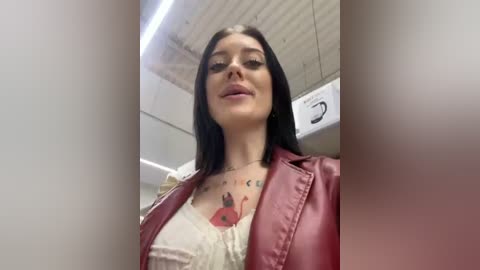 Media: Video of a young woman with long black hair, fair skin, and a tattoo on her chest. She wears a red leather jacket over a white shirt. The background shows an airplane cabin with overhead compartments.