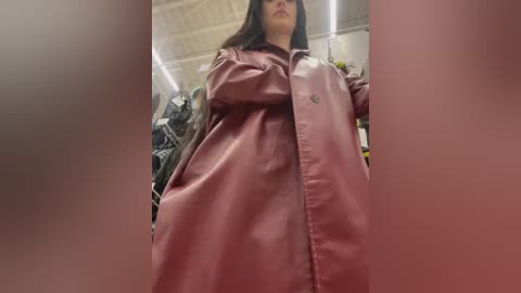 Media: Video of a woman in a long, maroon raincoat, standing in an industrial setting with fluorescent lighting and a cluttered background.