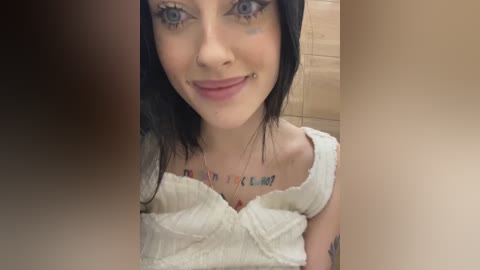 Media: Video of a young woman with pale skin, blue eyes, and black hair. She wears a white off-shoulder top and has a tattoo with words on her chest. Background features beige tiles.