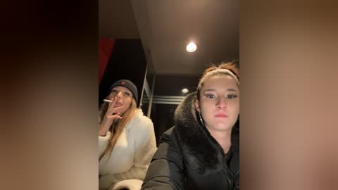 Media: Video of two young women, one smoking a cigarette, the other with a fur-lined jacket, in a dimly lit room with a recessed light fixture.