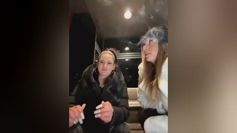 Media: Video of two women in a dimly lit room, one with long brown hair, the other with blonde hair, both smoking cigarettes, wearing fur coats, and sitting on a leather couch.