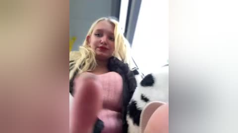 Media: A video of a blonde woman with large breasts and fair skin, wearing a black and white cow-patterned fur coat, standing in a brightly lit room with a window.