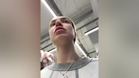 Media: A video of a young woman with fair skin, light brown hair, wearing a grey sweater, looking up, indoors with a modern ceiling and fluorescent lights.