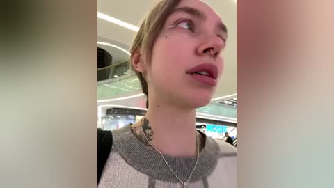 Media: Video of a young, fair-skinned woman with short blonde hair, wearing a grey and white sweater, standing in an airport. She has a tattoo on her neck and a silver necklace.