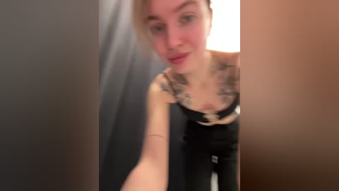 Media: A blurry video of a tattooed woman in black clothing, standing in a dark room, partially obscured by a vertical light-colored wall.