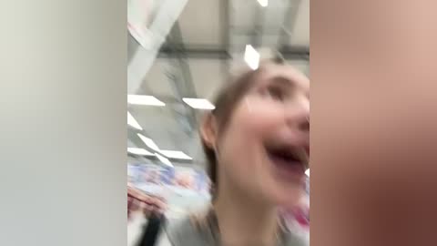 Media: A video of a young woman with light skin, brown hair, and a light gray top, smiling excitedly. The background features a blurred, modern indoor setting with large windows and bright lights.