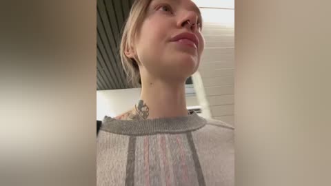 Media: Video of a young, fair-skinned woman with blonde hair, wearing a gray sweater with a colorful pattern, standing indoors against a white ceiling.