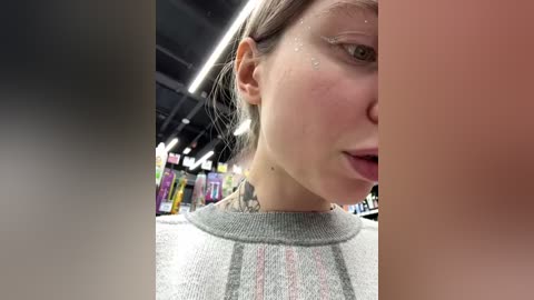 Media: Video of a young woman with pale skin, wearing a gray sweater, in a store with colorful products. Her face is partially blurred, revealing wetness and freckles.