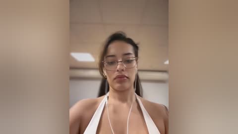 Media: Video of a young woman with medium skin tone, glasses, and a white halter top, seen from a low angle through a narrow gap, in a neutral indoor setting with beige walls and a ceiling.