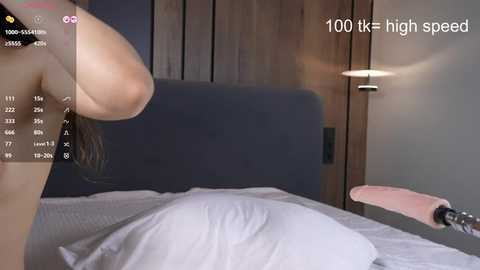 Media: Video of a woman with fair skin, long brown hair, and large breasts, wearing a black top, in a modern bedroom with a wooden headboard, white bedding, and a pink cushion on a bench.