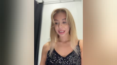 Media: Video of a blonde woman with straight hair, wearing a black and white floral-patterned dress, smiling with braces, standing in a bathroom with a shower curtain and white walls.