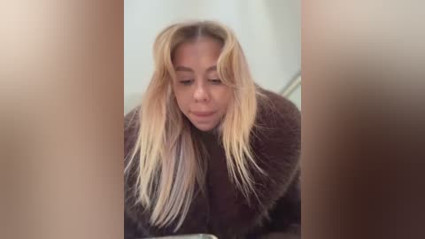 Media: Video of a woman with long, wavy blonde hair, wearing a dark brown fur coat, leaning forward with a playful expression, partially obscured by a blurred, light-colored background.