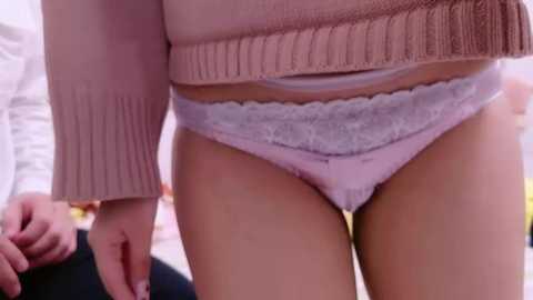 Media: Video of a woman's lower torso in light pink lace panties, partially visible white bra, and a beige sweater. Background shows blurred papers and a blurred person in the foreground.