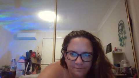 Media: A video of a middle-aged woman with glasses, fair skin, and brown hair, sitting topless at a cluttered vanity in a dimly lit bedroom.