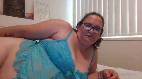 Media: A plus-sized woman with glasses, wearing turquoise lace lingerie, lies on a bed in a bedroom with white blinds and a framed picture.