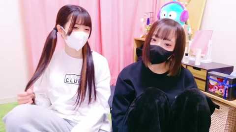 Video of two young Asian women with long black hair, wearing white masks and gray sweatshirts. One sits, the other stands in a cozy, pink-draped room with toys and a shelf.