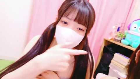 Media: Video of an East Asian woman with long dark hair and blunt bangs, wearing a white mask, pointing at the camera, in a pink room with a stuffed animal on a shelf.