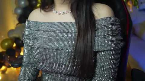 Media: Video of a woman with long, straight black hair, wearing an off-shoulder, sequined silver dress with a delicate necklace. Background features a blurred festive setting with golden and silver balloons.