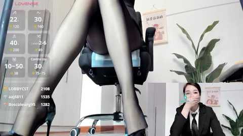 Media: Video of a woman in a black suit, wearing sheer black tights, sitting on a chair, in a modern office with a calendar, plants, and a desk.