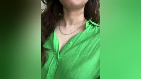 Media: Video of a woman with light skin, long brown hair, wearing a bright green, button-up shirt with a single button undone, revealing a hint of cleavage, and a delicate gold necklace. The background is a blurred gradient of green.