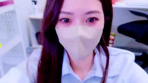 Media: Video of an East Asian woman with long, straight dark hair, wearing a light blue shirt and a beige face mask, indoors in a modern office setting with white walls and furniture.