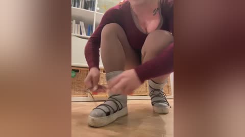 Media: A video captures a woman crouching, adjusting her gray sneakers on a light wooden floor. She wears a maroon top and beige shorts, with a tattoo on her neck. The background features a white shelf and a wicker basket.