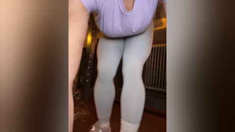 Media: A video of a person wearing light purple top and light blue leggings, bending over, with blurred background including a radiator and door frame.