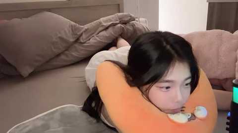 Media: Video of an Asian woman with long black hair, lying on a bed with a gray blanket, wearing a white shirt. She rests her head on an orange pillow with a white frog plushie. Background shows pillows and a beige wall.