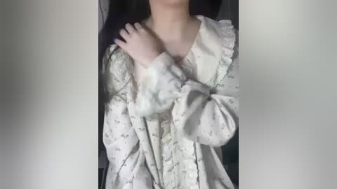 Media: Video of a person with fair skin, wearing a loose, off-white floral-patterned blouse with long sleeves, standing against a blurred, neutral background.