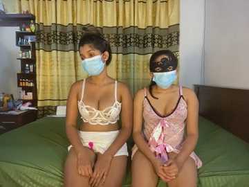 Media: Video of two women in matching white lace lingerie, blue masks, and face shields, sitting on a green bed, in a modestly decorated room with beige curtains and a dark wooden headboard.