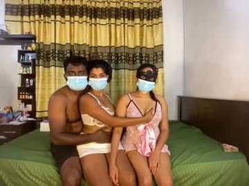 Media: Video of a muscular Black man, a slender Indian woman, and a petite Indian woman in face masks, sitting on a green bed in a simple bedroom with yellow curtains.