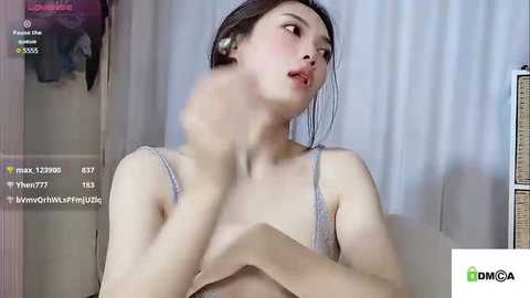 Media: Video of a fair-skinned, Asian woman with dark hair, wearing a sheer, light pink bra, in a living room. She's holding her bra straps with a confused expression.