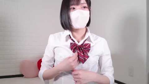 Media: Video of an East Asian woman with short black hair, wearing a white shirt, red plaid bowtie, and a white face mask, adjusting her shirt collar. Background features a white brick wall and a pink cushion.