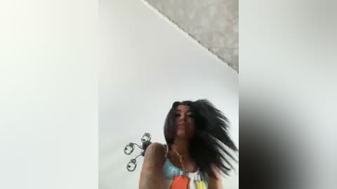 Media: Video of a woman with long black hair in a blue tank top, looking down at a black wall-mounted fan. Background includes a white ceiling and textured grey wall.