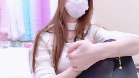 Video of a woman with long brown hair, wearing a white mask, white knit top, and black pants, seated in a room with pastel-colored curtains and a white bed.