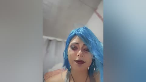 Media: Video of a woman with bright blue, shoulder-length hair and dark makeup, leaning against a white wall. She wears a black top and has a neutral expression.