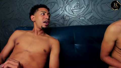 Media: Video of a shirtless, muscular, Black man with short hair and a goatee, sitting on a blue leather couch. He looks surprised, with a grey, textured wall behind him.