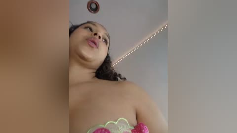 Media: A low-angle video of a curvy, light-skinned woman with curly hair, wearing a strapless pink and green lace top, captured from a slightly tilted angle. The background is a plain, light-colored wall with a small, circular light fixture.