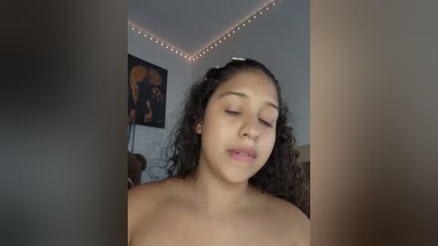 Media: A video of a young, topless woman with medium brown skin and curly hair, eyes closed, standing in a softly lit room with a framed picture and string lights on the ceiling.