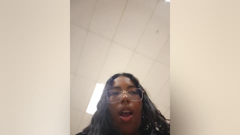 Media: Video of a young Black woman with medium-brown skin and long, wavy black hair, wearing glasses and a dark jacket, captured from a low angle, indoors with a white ceiling and fluorescent lights.