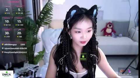 Media: Video of an Asian woman with long black hair, wearing a black cat ear headband, sitting in a modern, white room with a plush teddy bear and plants.