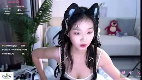 Media: Video of a young East Asian woman with long black hair, wearing a white bra and black cat ears, in a modern living room with a plush toy and green plant.