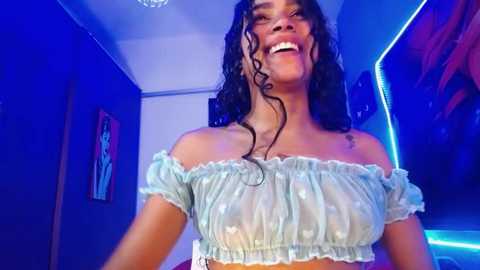 Media: Video of a joyful, medium-dark-skinned woman with curly hair in a light blue, see-through off-shoulder top, standing in a dimly-lit room with blue lighting and art on the walls.