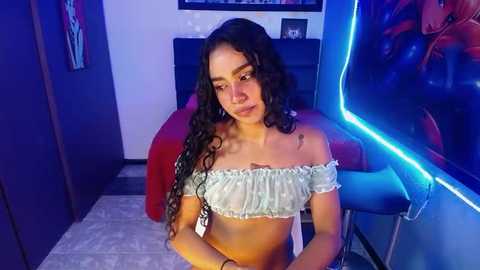 Media: Video of a young woman with long, curly black hair, wearing a light blue off-shoulder crop top, seated in a dimly lit room with neon blue lights and a red chair.