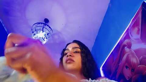 Media: Video of a young woman with curly black hair, medium skin tone, and full lips, standing in a brightly lit room with blue and purple accents, holding a cigarette.