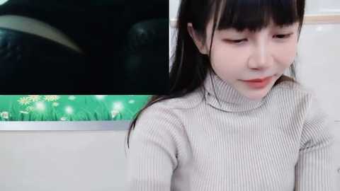 Media: A video of an Asian woman with straight black hair, wearing a beige ribbed turtleneck sweater, looking down thoughtfully. Background shows a green abstract wall art with white flowers and a black car window.