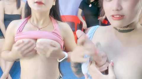 Media: Video of two young Asian women with light skin, one lifting her pink top to expose her small breasts, the other covering her nipples with her hand, surrounded by a casual outdoor crowd.