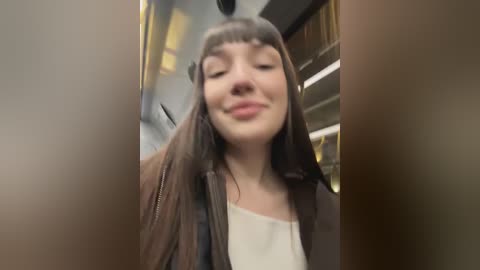 Media: A video of a young woman with long, straight brown hair, wearing a white top and brown jacket, smiling broadly. The background shows a blurred interior with shelves and yellow objects.