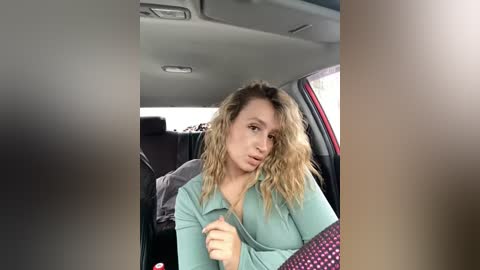 Media: A video of a blonde woman with wavy hair in a teal blouse, seated in a car, with blurred surroundings.