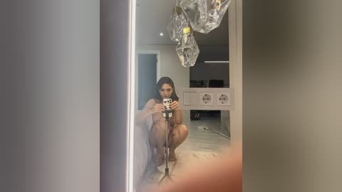 Media: Video of a nude woman with long dark hair crouching, taking a selfie in a modern bathroom with a clear glass ceiling light fixture, gray walls, and white storage units.